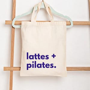 Puede incluir: Bolso tote de color crema con las palabras "lattes + pilates." impresas en azul oscuro. La bolsa est&aacute; colgada de una percha verde claro, que descansa sobre una escalera de madera.