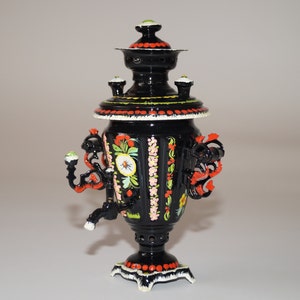 Może przedstawiać: Czarny, bogato zdobiony samowar z kwiatowymi i liściastymi wzorami w kolorze czerwonym, żółtym i zielonym. Samowar posiada kran, uchwyty i dekoracyjne zwieńczenie. Podstawa i górna krawędź są białe z czerwonymi akcentami.