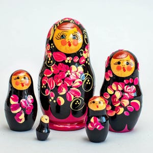 May include: A set of five Russian nesting dolls, or Matryoshka dolls, in varying sizes. The dolls are painted black with floral designs in pink and yellow. Each doll has a painted face with rosy cheeks and a red mouth.