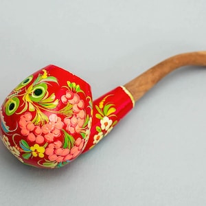 Op de afbeelding: Een handbeschilderde tabakspijp met een rode ketel en een lichtbruine steel. De ketel is versierd met roze, gele en groene bloempatronen. De steel is gebogen en van hout.