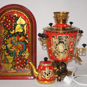 Puede incluir: Un samovar ruso rojo, dorado y negro con una tetera y una bandeja a juego. El samovar y la tetera tienen diseños florales. La bandeja presenta una colorida pintura de arte popular de un pájaro y bayas. El conjunto es de estilo tradicional.
