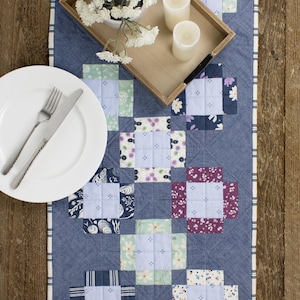 Peut inclure: Chemin de table matelassé bleu avec un motif patchwork composé de carrés aux motifs floraux et géométriques variés. Deux assiettes blanches avec couverts, un plateau en bois avec bougies et fleurs, sont disposés sur le chemin de table, sur une table en bois.