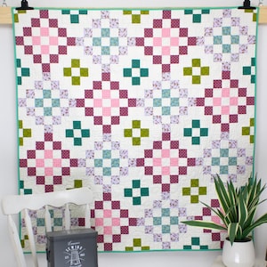 Op de afbeelding: Een handgemaakte quilt met een herhalend patroon van roze, groene, teal en paarse vierkanten op een witte achtergrond. De quilt wordt aan een muur getoond. Een witte stoel, een kleine bijzettafel en een plant in een witte pot staan op de voorgrond.