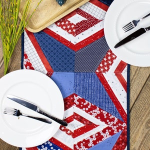May include: A red, white, and blue quilted table runner with a geometric pattern. Two white plates with silver cutlery are placed on the runner, along with a wooden tray with a small blue bird figurine and a vase with white flowers.