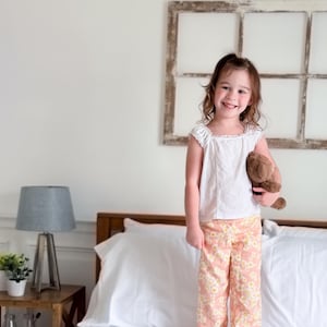 May include: A white sleeveless top with a square neckline and peach-colored pants featuring a white daisy floral pattern. The pants have a relaxed fit and the top has subtle vertical stitching details. A brown plush teddy bear is held in one hand.
