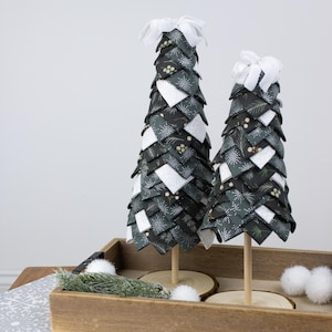 以下が含まれることがあります： ダークグリーンと白の模様の装飾用布製クリスマスツリー2本。白いリボンが飾られています。木製のダボの上にあり、装飾的なアクセントが付いた木製トレイに置かれています。ツリーの高さは約25cmと20cmです。