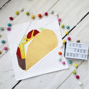 May include: A paper pieced quilt block featuring a yellow and brown taco with red and yellow fillings. The block is on a white background with a white light box that says "Lets Taco Bout It".