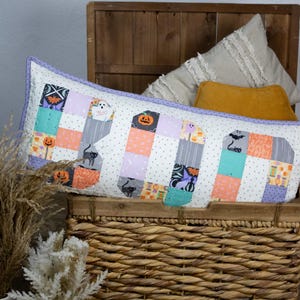 May include: A rectangular Halloween-themed pillow with the word "BOO" appliqued on the front. The letters are made of patchwork squares with Halloween motifs like pumpkins, bats, and ghosts. The pillow has a white background with small gray dots and a lavender border.