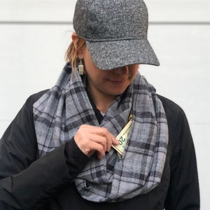 Puede incluir: Una bufanda infinita de cuadros grises y negros con un bolsillo con cremallera. El bolsillo está abierto y se ve un billete de dólar.