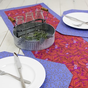 Pode incluir: Um corredor de mesa vermelho e roxo com um padrão floral e um porta-garrafas de leite de metal com três garrafas de vidro. O corredor de mesa é rodeado por placemats azuis com um padrão floral branco.