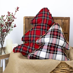 May include: Two Christmas tree-shaped cushions with frayed edges. One is red with a green and black plaid pattern, and the other is white with a red, black, and grey plaid pattern. They are displayed in a woven basket with a burlap liner, suggesting a festive holiday theme.