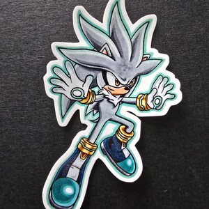 May include: A sticker featuring Silver the Hedgehog in a dynamic pose. The character has gray fur, a white chest, and gold accents on his wrists and shoes. His shoes are blue with teal details. The sticker has a white border and a teal outline.