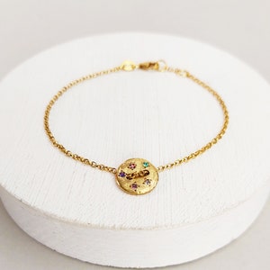 May include: A gold chain bracelet with a round charm featuring a textured surface and colourful gemstones.