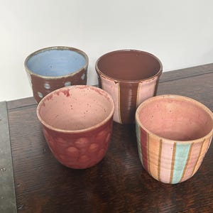 May include: Four handmade ceramic bowls in shades of pink, brown, and blue. One bowl has a light blue interior and brown exterior with white dots. Another has a pink interior and brown exterior with vertical stripes. The bowls are on a wooden surface.