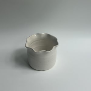 May include: A small, off-white ceramic vase with a ruffled rim. The speckled surface adds texture to the handmade pottery. The vase has a rounded body and a wide opening, suitable for small floral arrangements or decorative use.