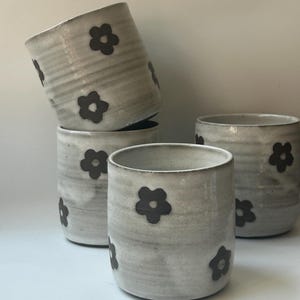 May include: A collection of handmade ceramic cups. The cups are a light gray color with a textured surface and dark brown flower designs. The cups are cylindrical in shape and appear to be of varying heights.