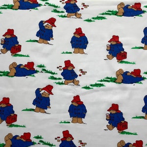 May include: White fabric with a repeating pattern of Paddington Bear in various poses. Paddington is wearing a blue coat, red hat, and carrying a brown suitcase with the initials "PB" on it.