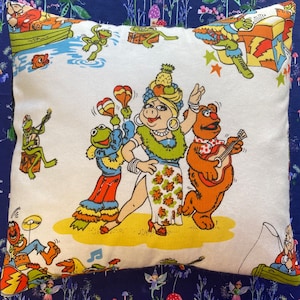 May include: Square cushion with a colourful cartoon scene featuring Miss Piggy, Kermit the Frog, Fozzie Bear, and other Muppets. The cushion has a white background with a vibrant, retro-style print.