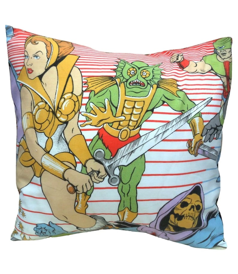 May include: Square pillow featuring a colorful cartoon illustration of action figures. The design includes a female warrior in gold armor, a green monster with a sword, and other characters on a white background with red stripes. The pillow is a decorative item.