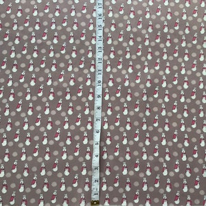 Puede incluir: Un primer plano de una tela marrón con un patrón repetido de conejos blancos que llevan abrigos rojos. La tela se mide con una regla en pulgadas.