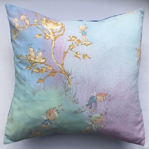May include: A blue and green cushion featuring a whimsical design of fairies and a bird's nest in a tree. The background is a soft watercolour wash of pink, blue, and green.