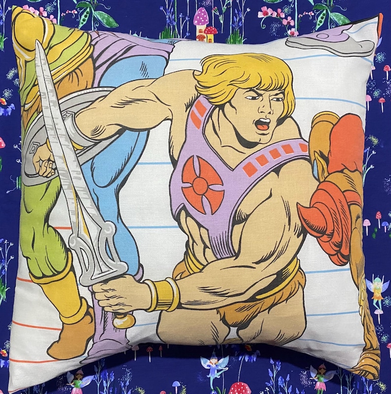 May include: Square pillow featuring a colorful illustration of a muscular cartoon character with blonde hair, wearing a purple chest plate and holding a sword. The pillow has a white background with blue lines and is set against a dark blue floral background.