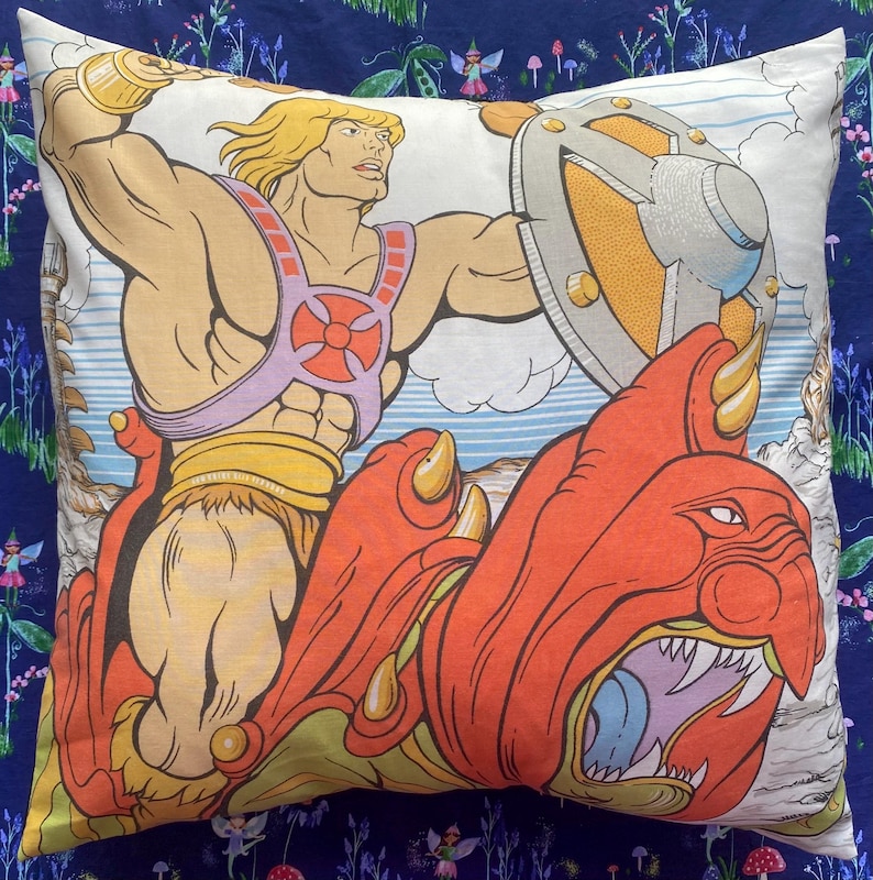 May include: Square pillow featuring a vintage illustration of a muscular blond character with a shield, riding a red creature. The pillow has a white background with a cartoon landscape and is bordered by a dark blue floral pattern.