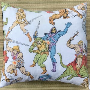 May include: White square pillow featuring a colorful action-packed illustration of various characters in a comic book style. The design includes figures with swords, shields, and other weapons, with a retro cartoon aesthetic.