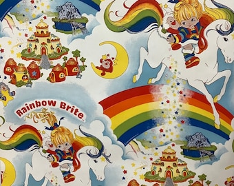 Puede incluir: Papel de regalo con temática de Rainbow Brite con un unicornio blanco con una crin y cola de arcoíris, una niña montando el unicornio y un castillo con detalles de arcoíris. El texto "Rainbow Brite" aparece dos veces en el papel.
