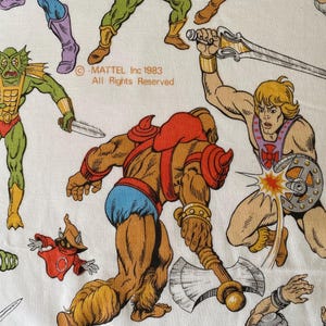 May include: White fabric featuring colorful illustrations of He-Man and other characters from the Masters of the Universe franchise. The image includes the text "MATTEL Inc 1983 All Rights Reserved."