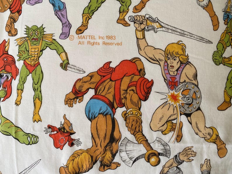 May include: White fabric featuring colorful illustrations of He-Man and other characters from the Masters of the Universe franchise. The image includes the text "MATTEL Inc 1983 All Rights Reserved."
