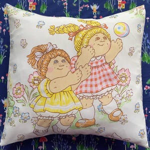 Peut inclure: Coussin carré décoratif avec une illustration vintage de deux enfants jouant avec une balle. Les enfants portent des robes et sont entourés de fleurs. Le coussin a un fond blanc et est bordé d'un motif floral bleu.