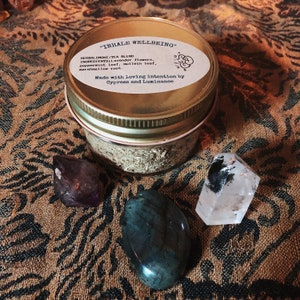 Puede incluir: Un frasco de vidrio con tapa dorada que contiene una mezcla de hierbas para fumar/té. La etiqueta dice "INHALE WELLBEING" y enumera los ingredientes: flores de lavanda, hojas de menta, hojas de gordolobo y raíz de malvavisco. El frasco está sobre una superficie marrón con estampado y tres cristales.