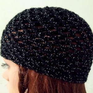 May include: A black crocheted beanie with silver metallic thread woven throughout. The hat has a textured, open weave design. The beanie is shown on a mannequin head with red hair.