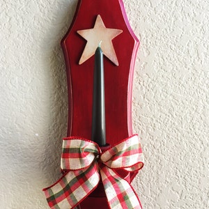 May include: A red wooden candle holder with a star cutout and a black candle. A plaid ribbon bow is tied at the base. The candle holder is designed to be mounted on a wall.