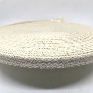 May include: A close-up of a roll of natural cotton webbing. The webbing is woven in a herringbone pattern and has a natural, off-white color.