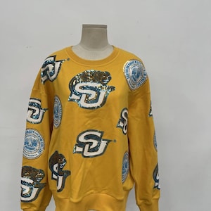 May include: Mustard yellow sweatshirt with multiple sequined "SU" logos and circular patches. The logos are in white and teal sequins. The sweatshirt is displayed on a mannequin.