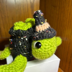 May include: A green crocheted turtle wearing a black and white witch hat with colorful accents. The turtle is sitting in a white box.