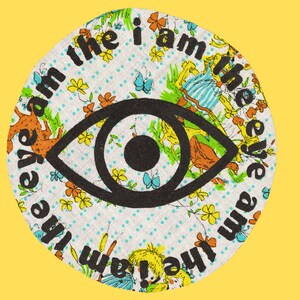 May include: A round, white fabric patch with a black eye graphic and the text "I am the eye am the eye am the eye" repeated around the edge. The fabric has a floral pattern with yellow, blue, and red flowers.