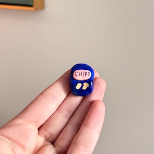 May include: A small, handmade blue sculpture of a chips packet with the word "CHIPS" in pink. Two chips are visible on the front. The sculpture is held in a hand against a neutral background.