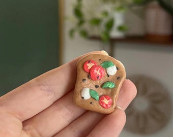 Puede incluir: Una pequeña escultura de arcilla marrón hecha a mano que representa una rebanada de pan cubierta con rodajas de tomate rojo, hojas de albahaca verde y queso mozzarella blanco.