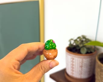 May include: A miniature hand-painted cactus figurine in a small brown pot. The cactus is green with pink flower accents. The figurine is held in a person's hand, with other potted plants visible in the background.