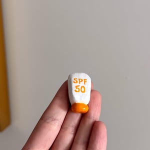 May include: A miniature white sunscreen bottle with orange accents. The text "SPF 50" is printed in orange on the front. The bottle is held in a hand.