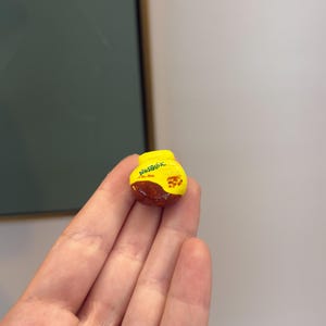 Puede incluir: Una pequeña roca pintada a mano con el tema de Nesquik. La parte superior es amarilla con la palabra "Nesquik" en verde. La mitad inferior es marrón. La roca está sostenida en la mano de una persona.