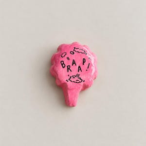 May include: A bright pink, cloud-shaped pin with a glossy finish. The center features the black text "BRAAP!" surrounded by small, hand-drawn cloud outlines. The object has a small, pointed extension at the bottom, resembling a speech bubble.