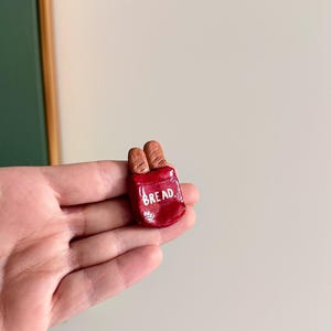 May include: A small, red ceramic bread bag with the word "BREAD" in white lettering. Two small, brown bread loaves are visible inside the bag. The item is held in a person's hand.