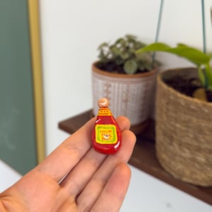 Op de afbeelding: Een kleine rode fles met een geel etiket met de tekst "Chili's BBQ". De fles heeft een lichtgekleurde stopper. De fles wordt in een hand gehouden, met planten op de achtergrond.