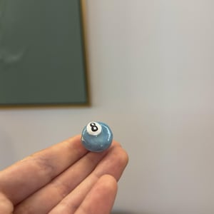 Op de afbeelding: Een kleine, lichtblauwe achtball met een witte cirkel en het nummer 8 in zwart. De bal is rond en lijkt gemaakt van een glanzend materiaal. De achtergrond is een effen witte muur.