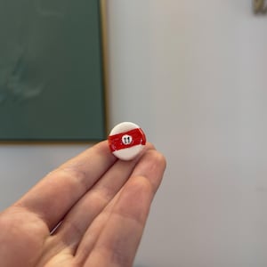 Könnte beinhalten: Eine kleine, runde, weiß-rote Billardkugel mit der Nummer 11. Die Kugel wird in einer Hand gehalten, mit einem unscharfen Hintergrund.