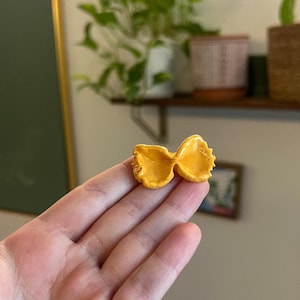 Pode incluir: Um único laço de massa de cerâmica amarelo. A massa tem a forma de um laço e é do tamanho de uma moeda de 20 cêntimos.
